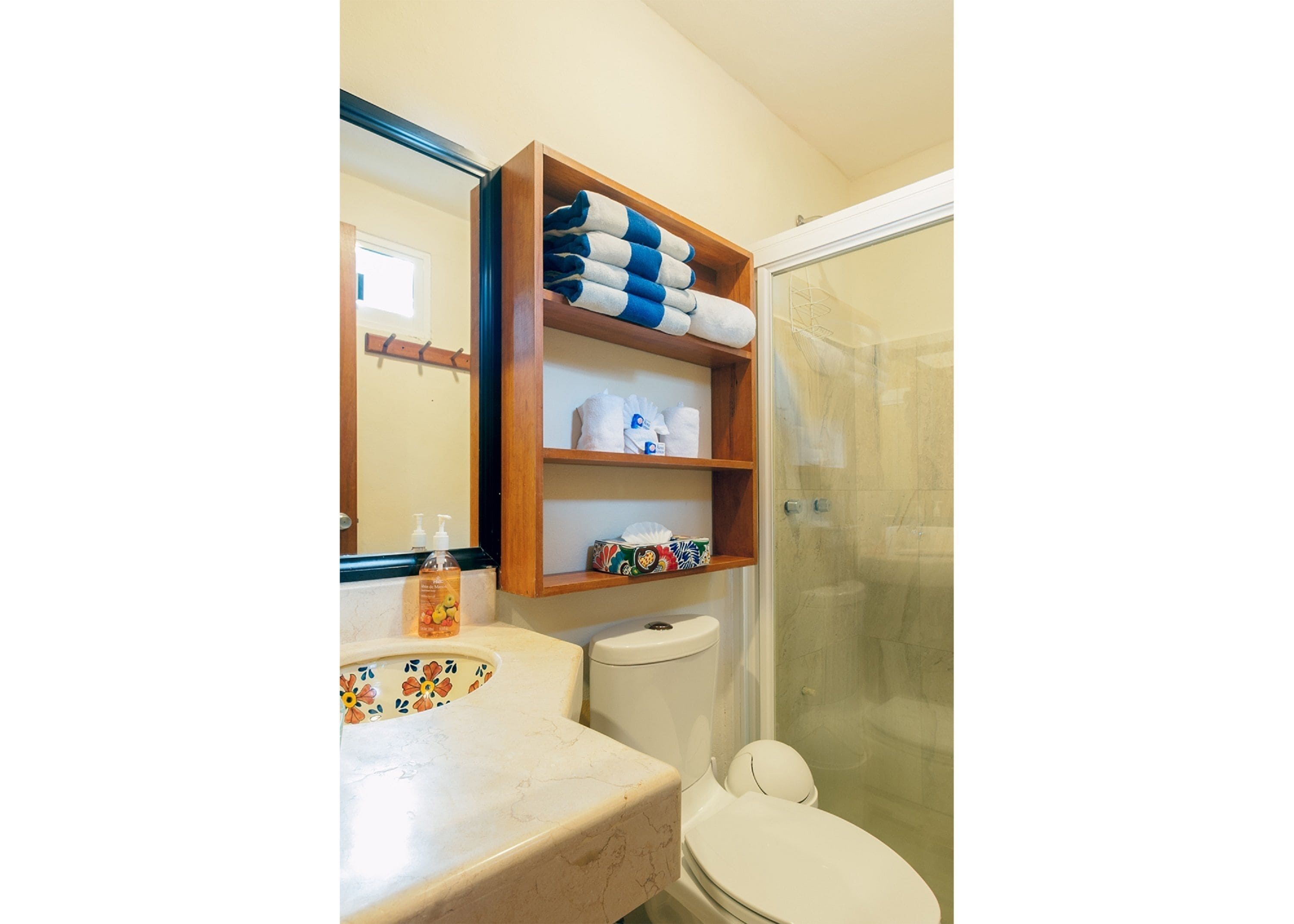 Ein Badezimmer mit Toilette, einem Waschbecken mit floralem Fliesendesign, einer Holzablage mit Handtüchern und einer Duschkabine aus Glas.