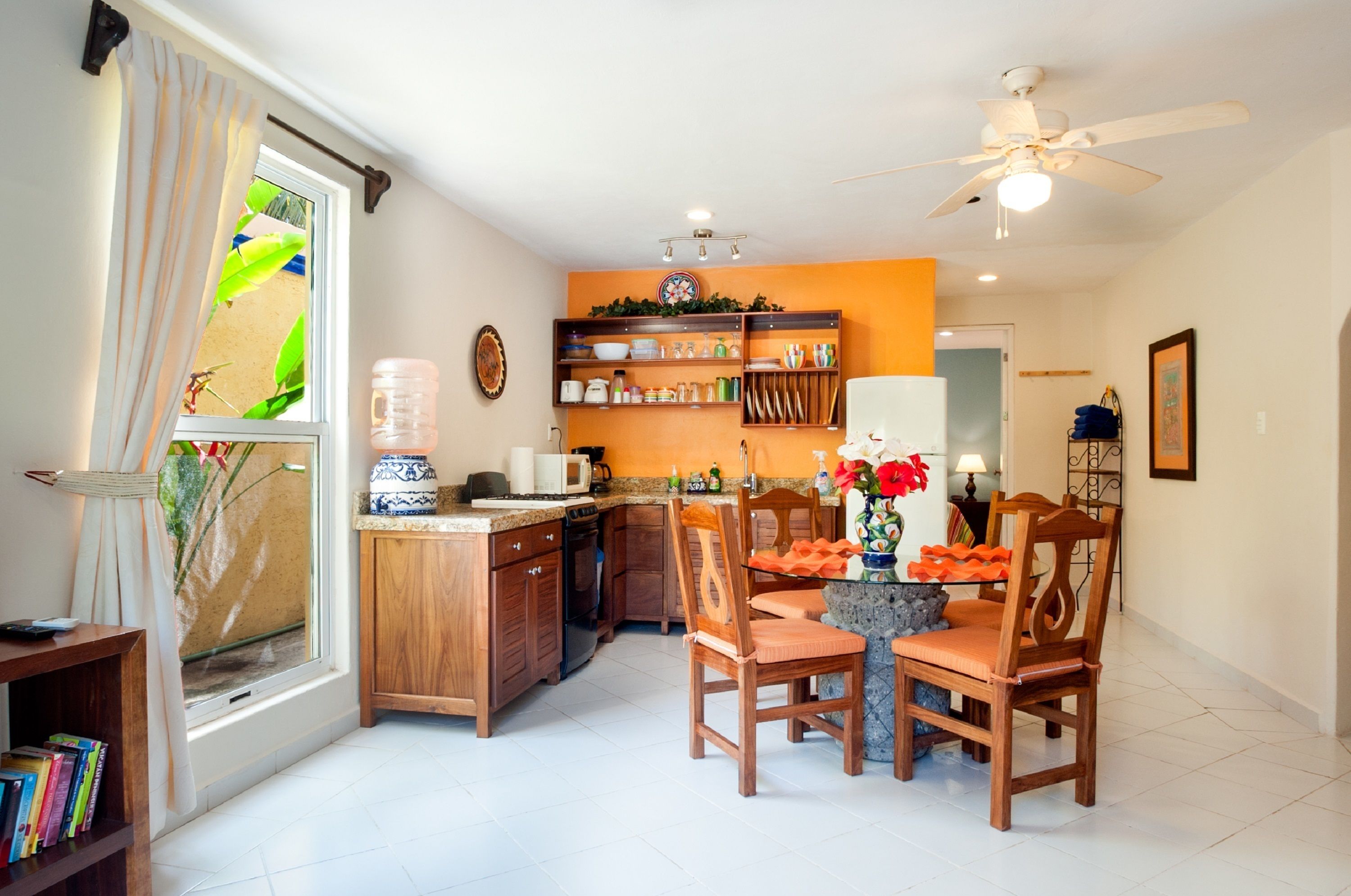 A modern kitchen with wooden cabinets, a dining area with two wooden chairs, a ceiling fan, and a window with curtains.