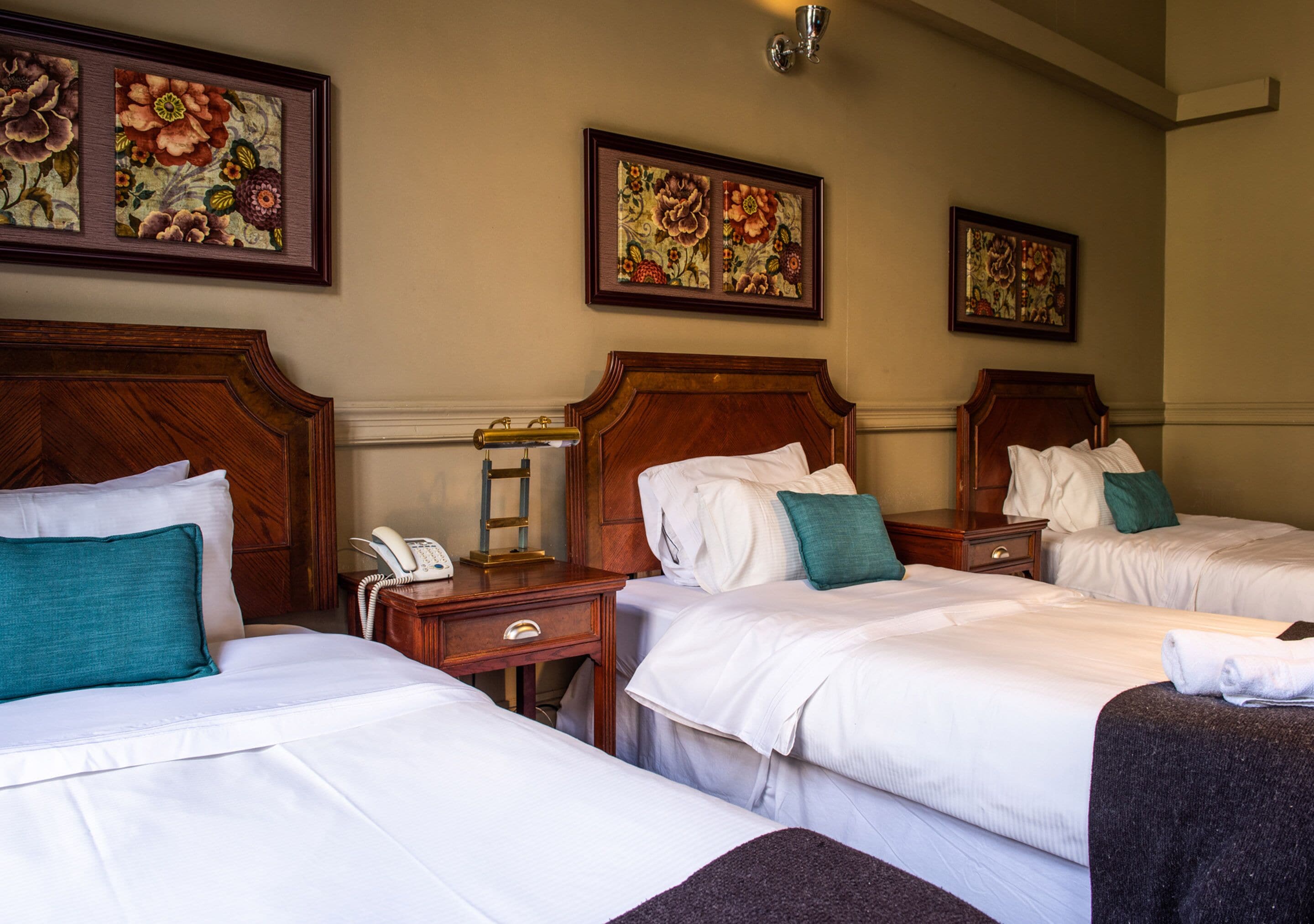 Ein Hotelzimmer mit drei Betten, Holz-Kopfteilen, weißen Laken und floralen Bildern an den Wänden.