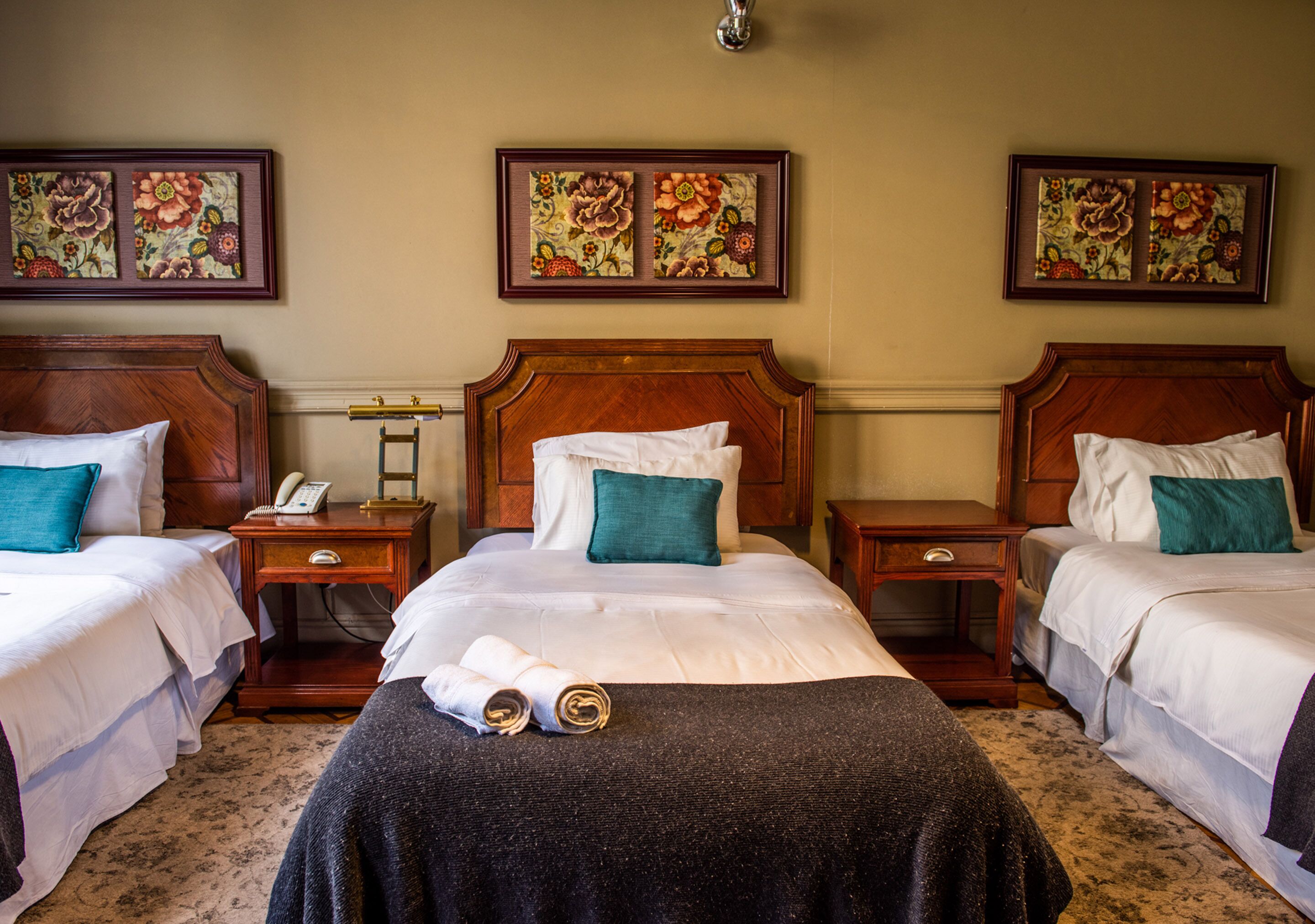 A hotel room with three beds, each with a wooden headboard, white bedding, and teal pillows. There are framed floral artworks on the wall above each bed.