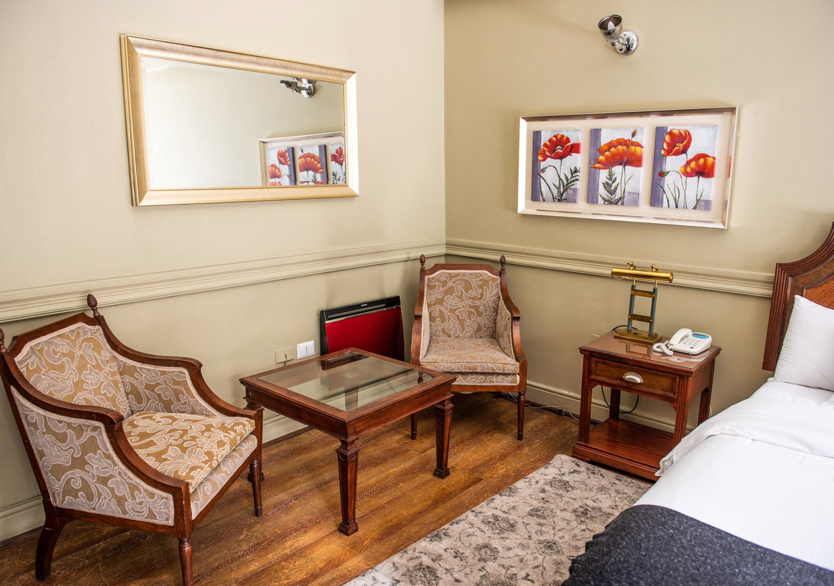 A hotel room with a bed, two armchairs, a small table, a nightstand with a lamp, a mirror, and framed artwork on the wall.