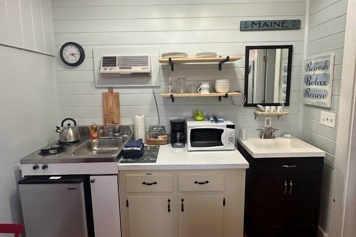 A compact kitchen with a sink, stove, microwave, and cabinets. There is a clock on the wall and a sign that says "MAINE".