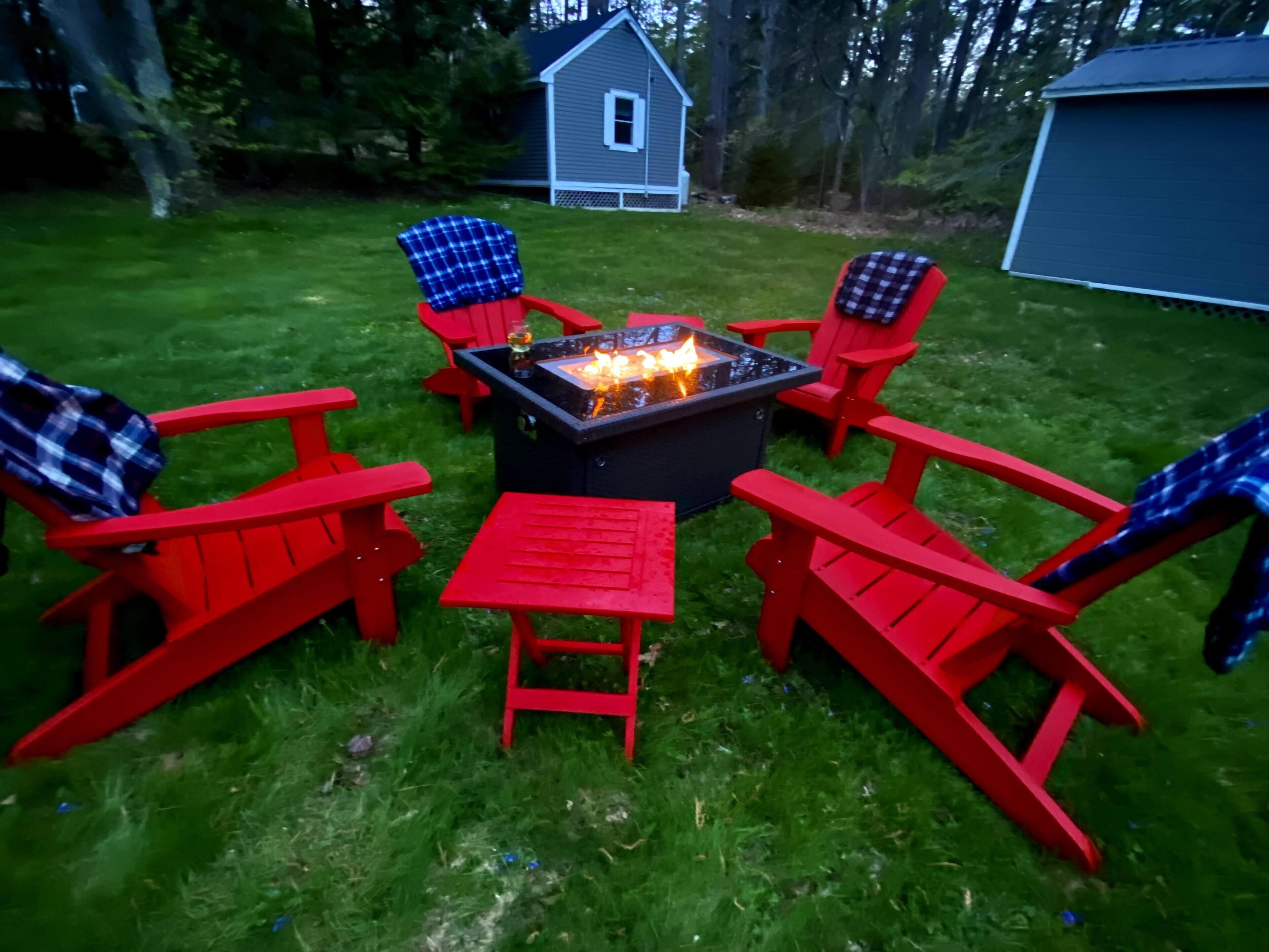 A fire pit surrounded by red Adirondack chairs and a small table in a grassy backyard with a shed and a house in the background.