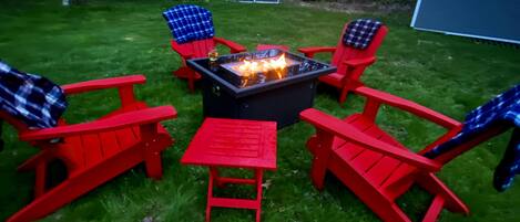 A fire pit surrounded by red Adirondack chairs and a small table in a grassy backyard with a shed and a house in the background.