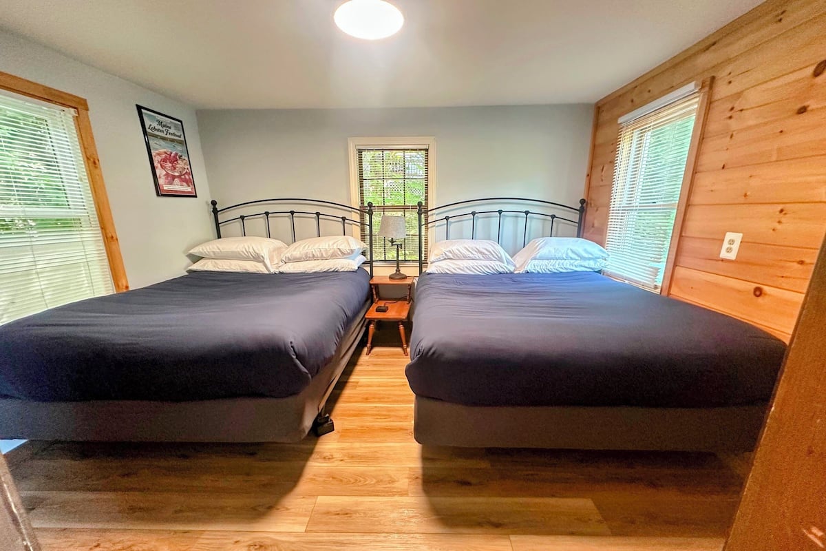 A room with two beds, a small table, a lamp, and a window with blinds.