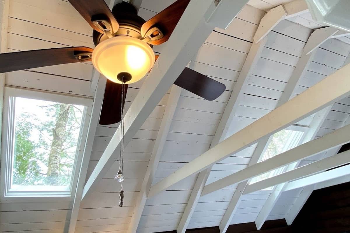 A ceiling fan with a light fixture in a room with white wooden beams and a window.