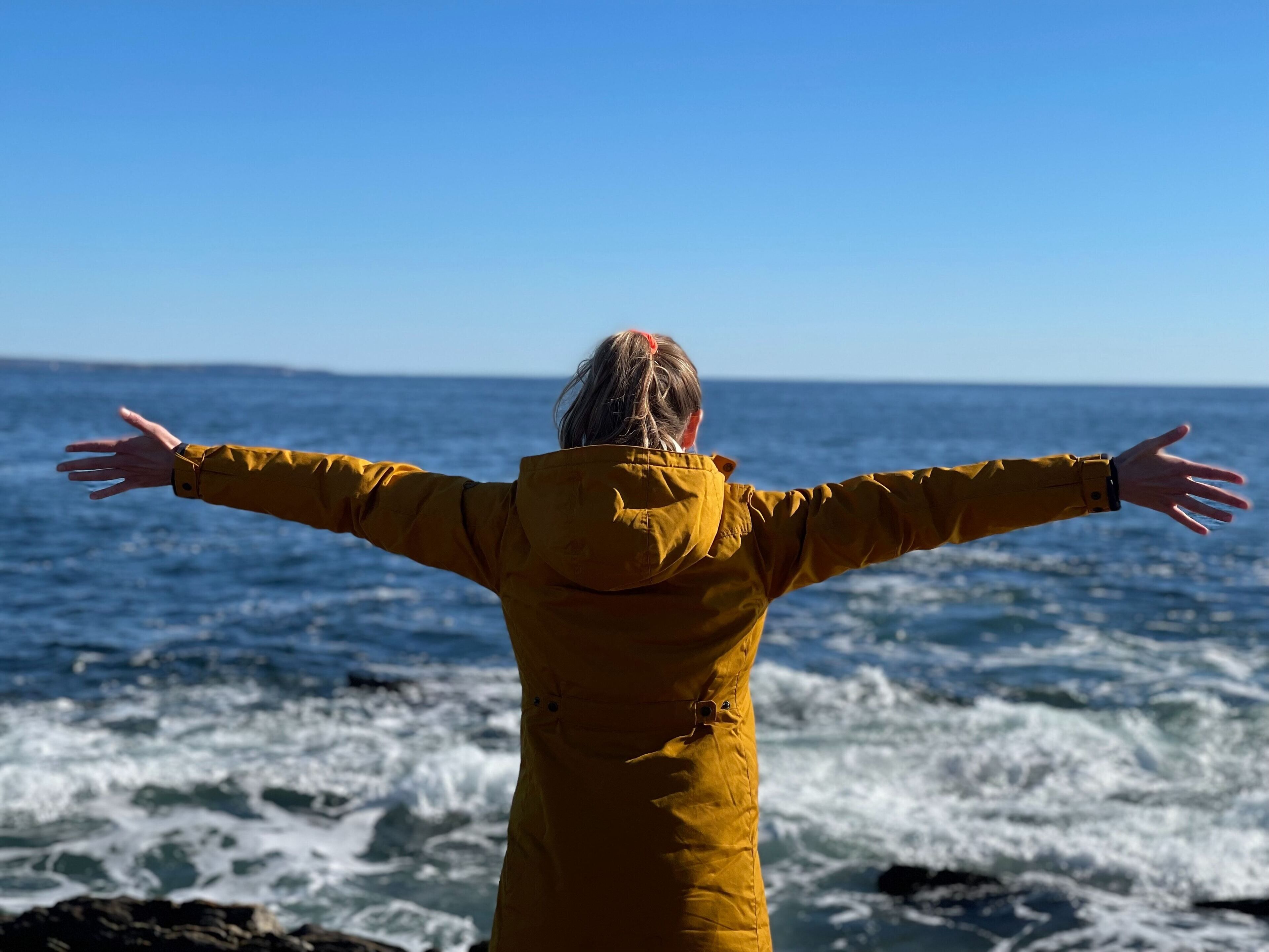 En person i en gul jakke med armene udstrakte, der ser ud over havet.