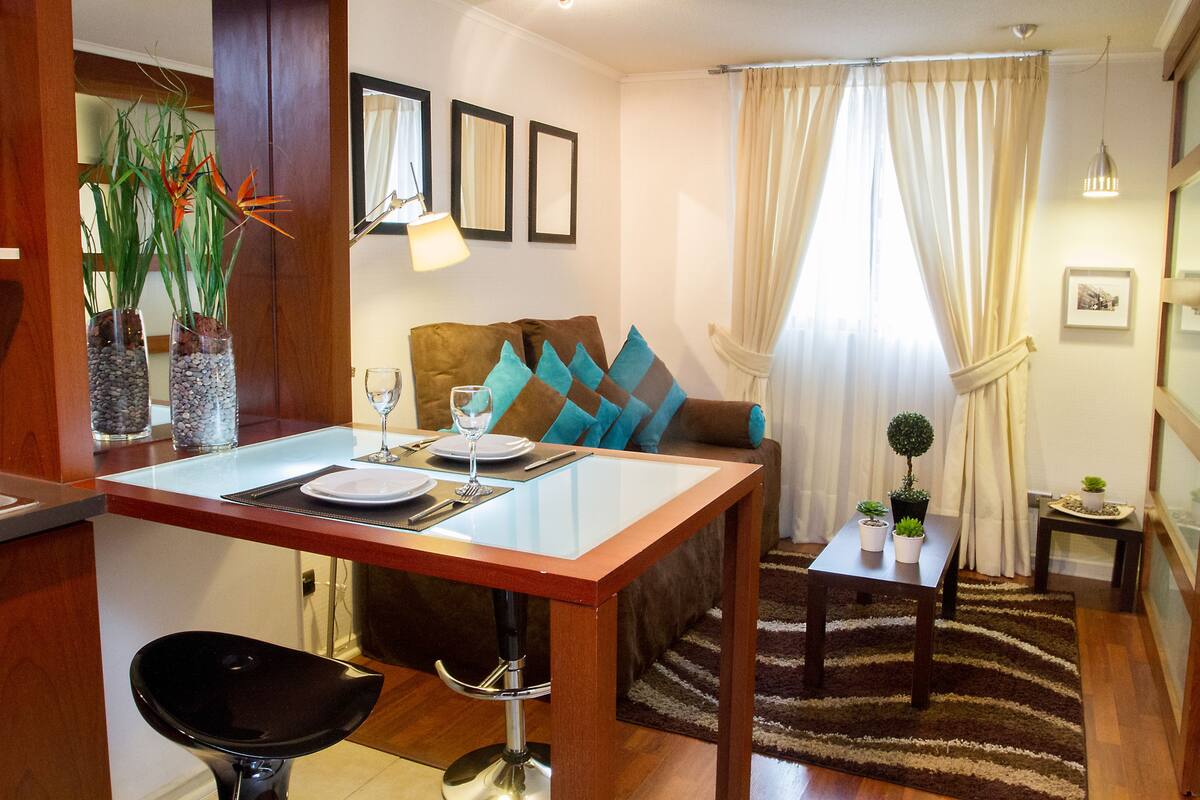 Uma área de refeições com uma mesa posta para dois, um sofá com almofadas, uma mesinha lateral com uma planta e vista da sala através de uma janela com cortinas.