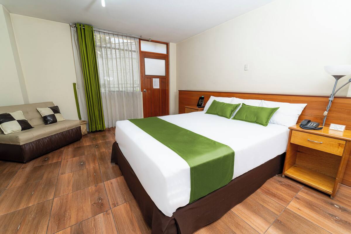 A hotel room with a bed, a wooden nightstand, a bedside table, a green and white bedspread, a brown sofa, and a window with green curtains.