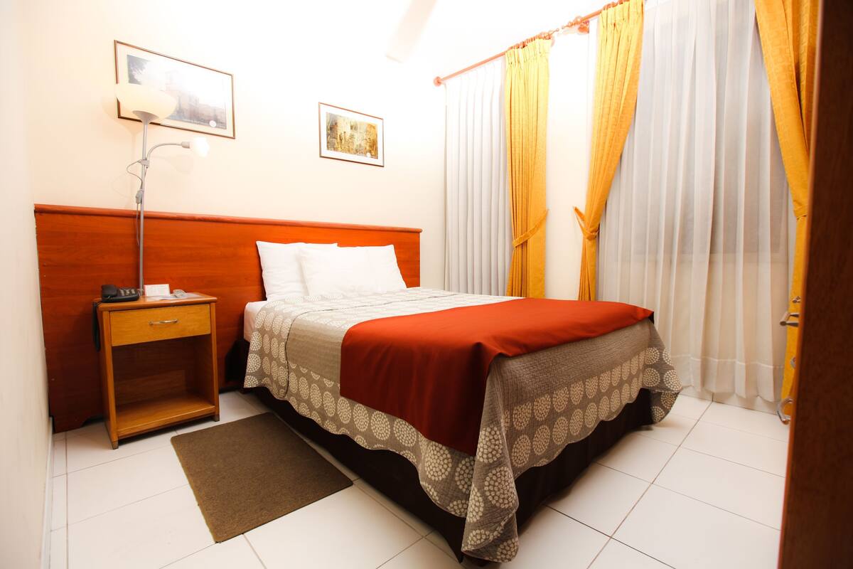 A hotel room with a wooden headboard, a single bed with a red and patterned bedspread, a bedside table with a lamp, and a window with yellow curtains.