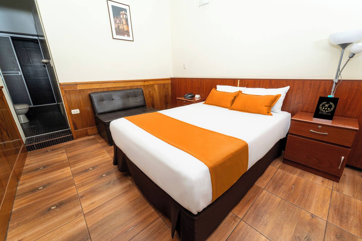A hotel room with a large bed, wooden headboard, a nightstand, a bedside table, and a framed picture on the wall.
