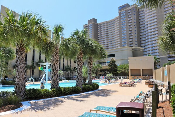 A hotel pool area with palm trees, lounge chairs, and a large building in the background.