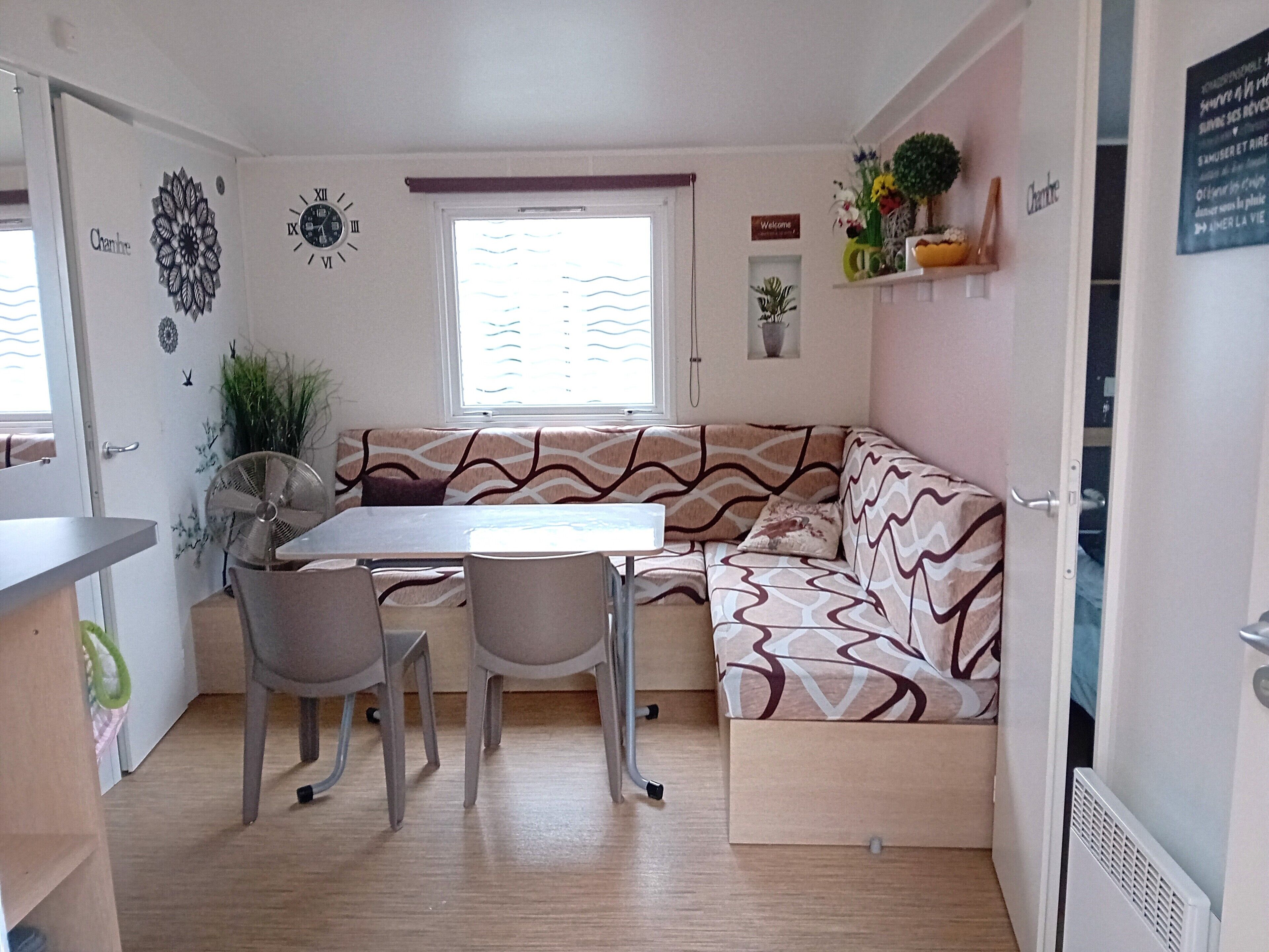 Ein kompakter Wohnraum mit Esstisch und Stühlen, einer Couch, einem Fenster mit Jalousien, einer Wanduhr und einem kleinen Regal mit Dekoration.