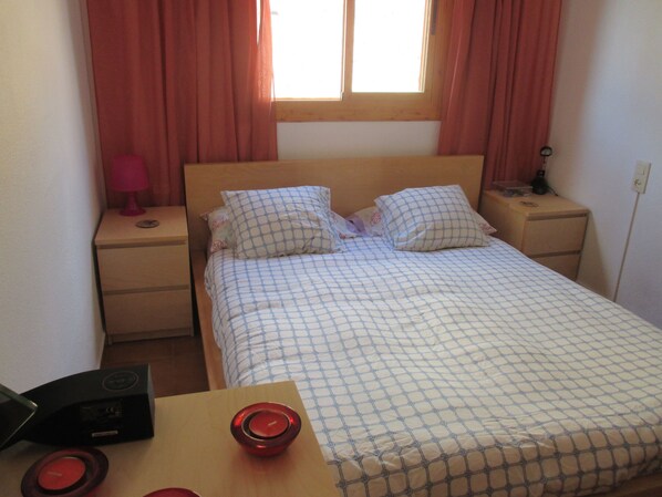 A small, neatly made bed with a checkered bedspread, two bedside tables with lamps, and a window with curtains.