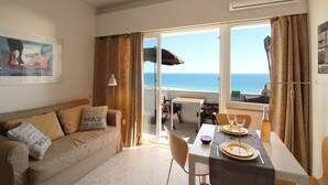 Une pièce comprenant un canapé, une table à manger dressée pour deux personnes et une vue sur la mer grâce à de grandes fenêtres.