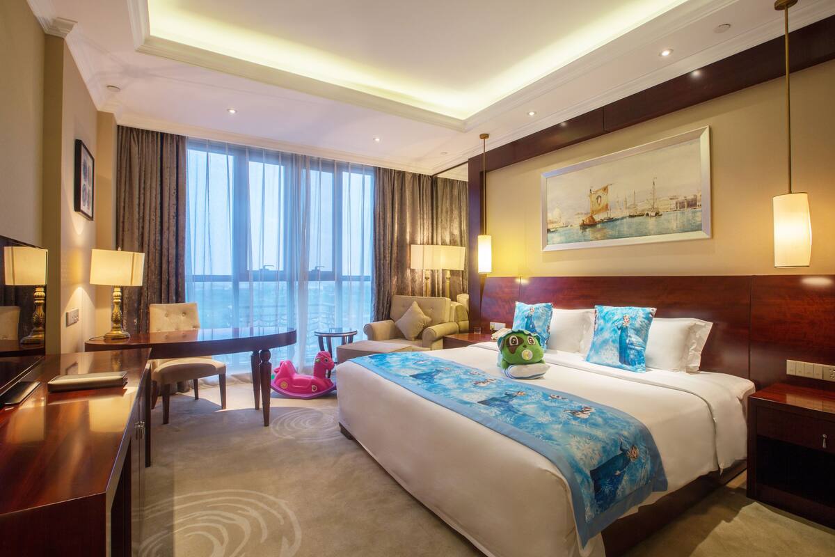 Ein Hotelzimmer mit einem großen Bett, einem Schreibtisch, einem Stuhl, einem kleinen Tisch und einem Fenster mit Vorhängen.