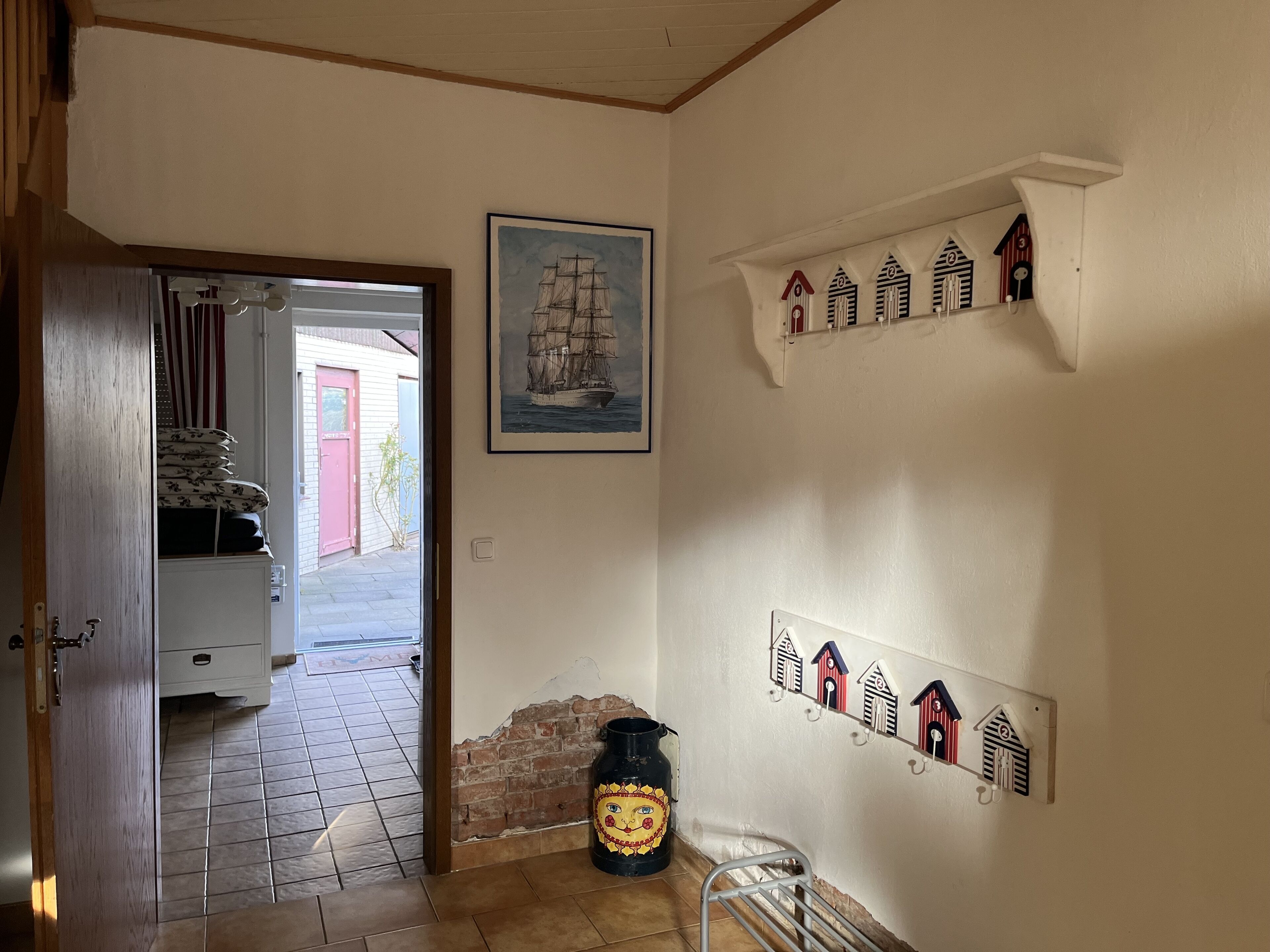 A hallway with a tiled floor, a door leading to an outdoor area, a framed picture of a ship, and wall-mounted hooks.