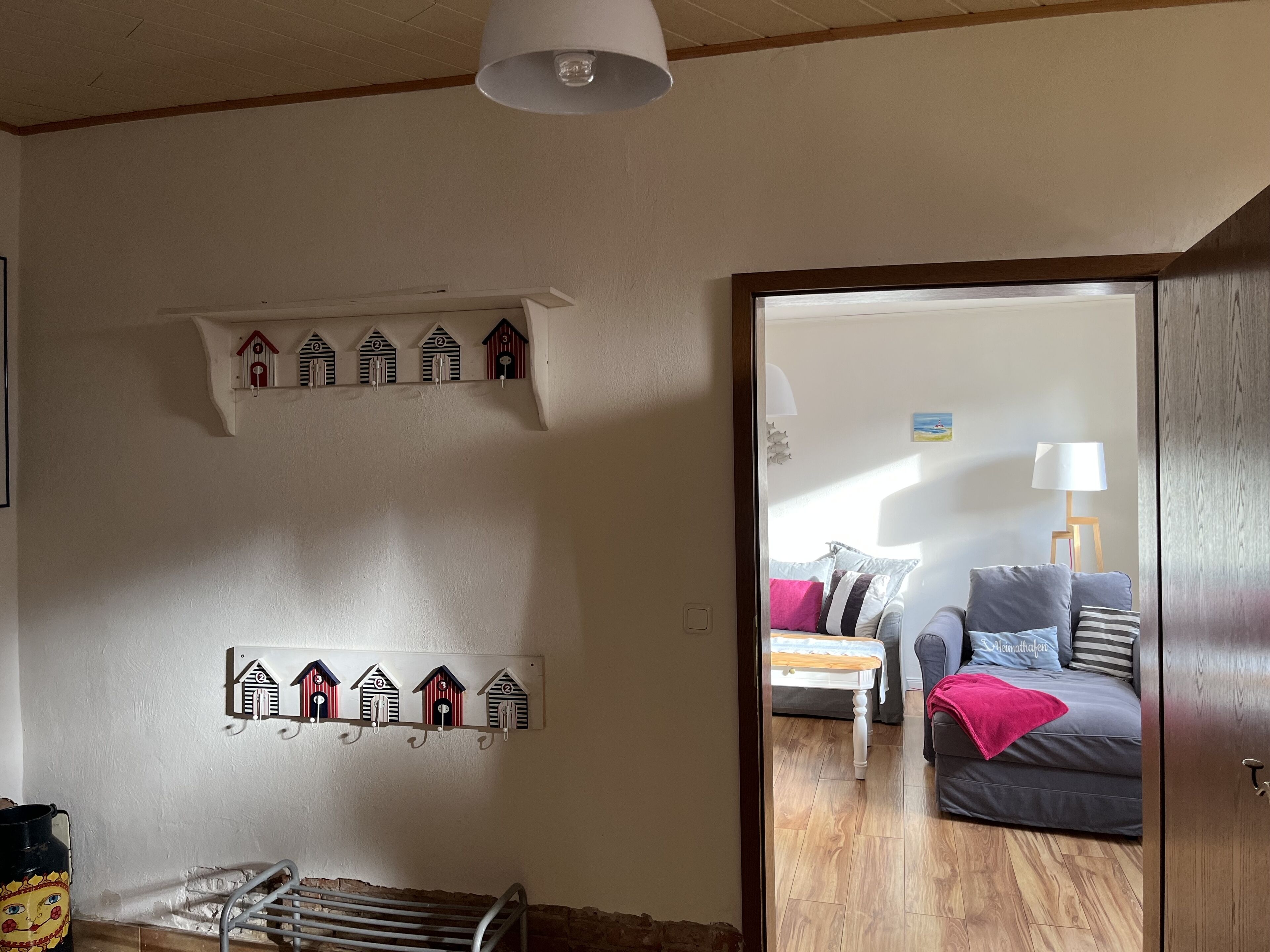 Une pièce dotée d’un plancher en bois, d’un mur blanc orné d’une rangée de décorations représentant des nichoirs, d’un banc en métal et d’une porte donnant sur une autre pièce où se trouvent un canapé et une lampe.