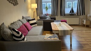 Una sala de estar moderna con un sofá gris, una mesa de centro de madera y vistas a un edificio a través de ventanas grandes con cortinas blanco y negro.