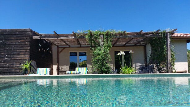 Een zwembad met helder water, een houten pergola en een loungehoek met gestreepte stoelen.