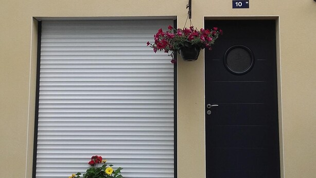 Un edifici amb una persiana metàllica abaixada, una porta negra amb una placa identificadora i una petita finestra amb jardineres florals penjants.