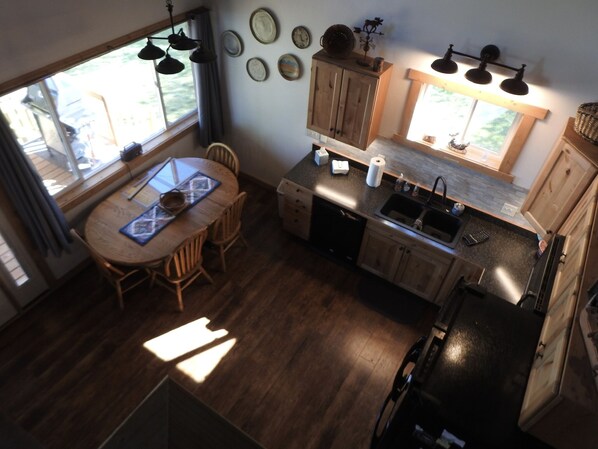 A compact kitchen with a dining table, wooden cabinets, a dishwasher, and a window providing natural light. - Duck Lake West Shore, Just 15 Minutes from Glacier National Park! (Babb)