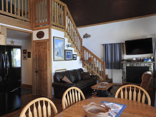 A cozy living room with a wooden staircase, a fireplace, a black refrigerator, a leather sofa, and a dining table with chairs. - Duck Lake West Shore, Just 15 Minutes from Glacier National Park! (Babb)