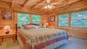 Ein Schlafzimmer einer Holzhütte mit einem Bett, zwei Fenstern, einem Deckenventilator und einem Nachttisch mit Lampe.