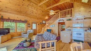 Ein Blockhaus-Interieur mit Holzvertäfelung, einer Treppe, einem Wohnzimmer mit Sofa, einem Fernseher und einer Küchenzeile.