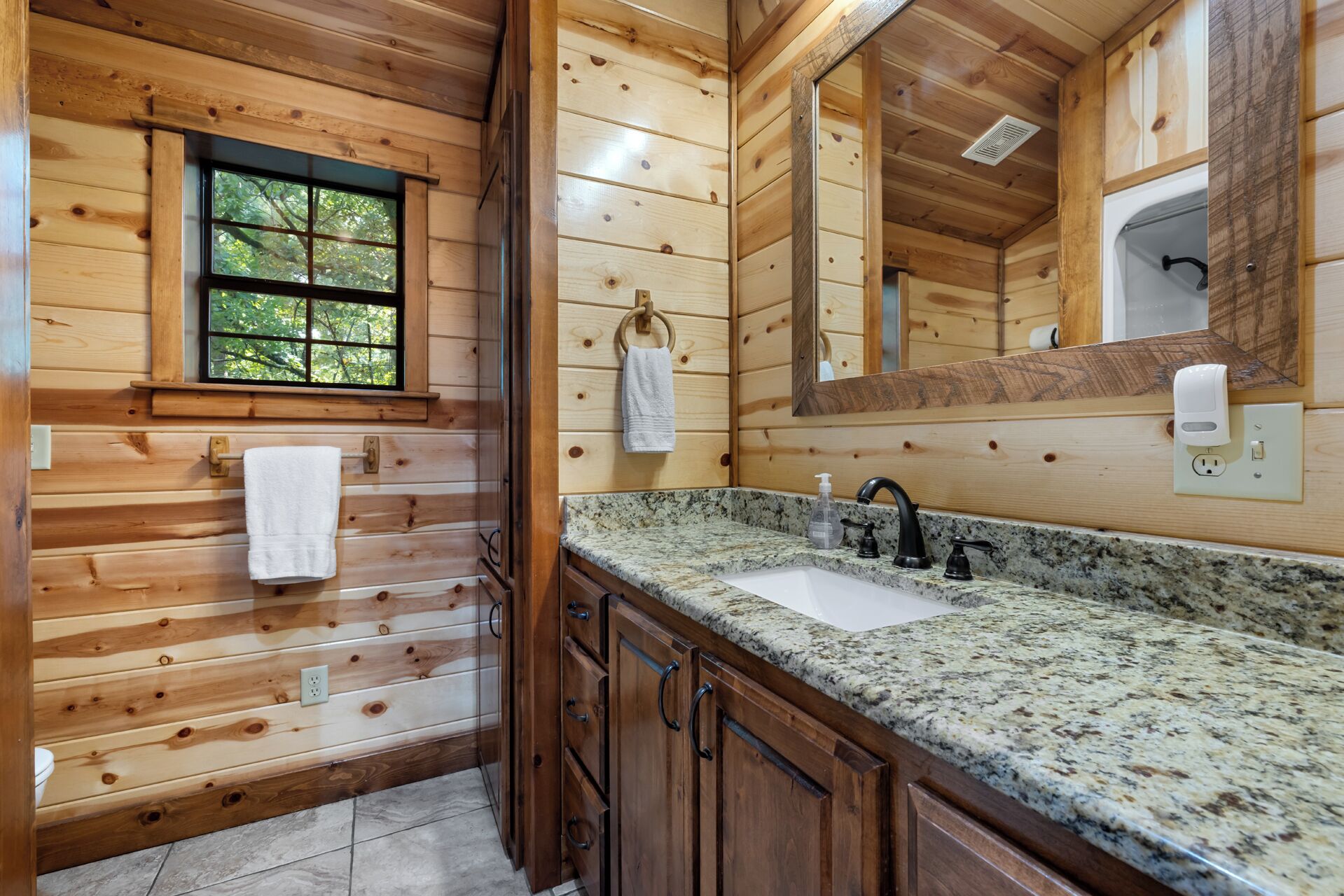 A wooden cabin bathroom with a granite countertop sink, wooden cabinets, a window, and a mirror.