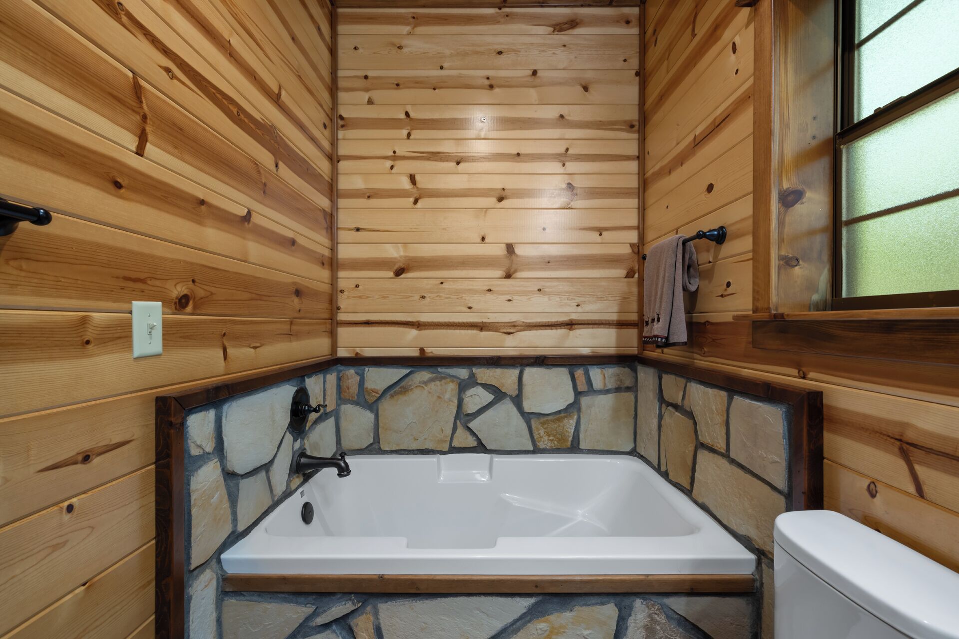 Baño de cabaña de madera con pared de mosaicos de piedra, una bañera blanca y un inodoro.
