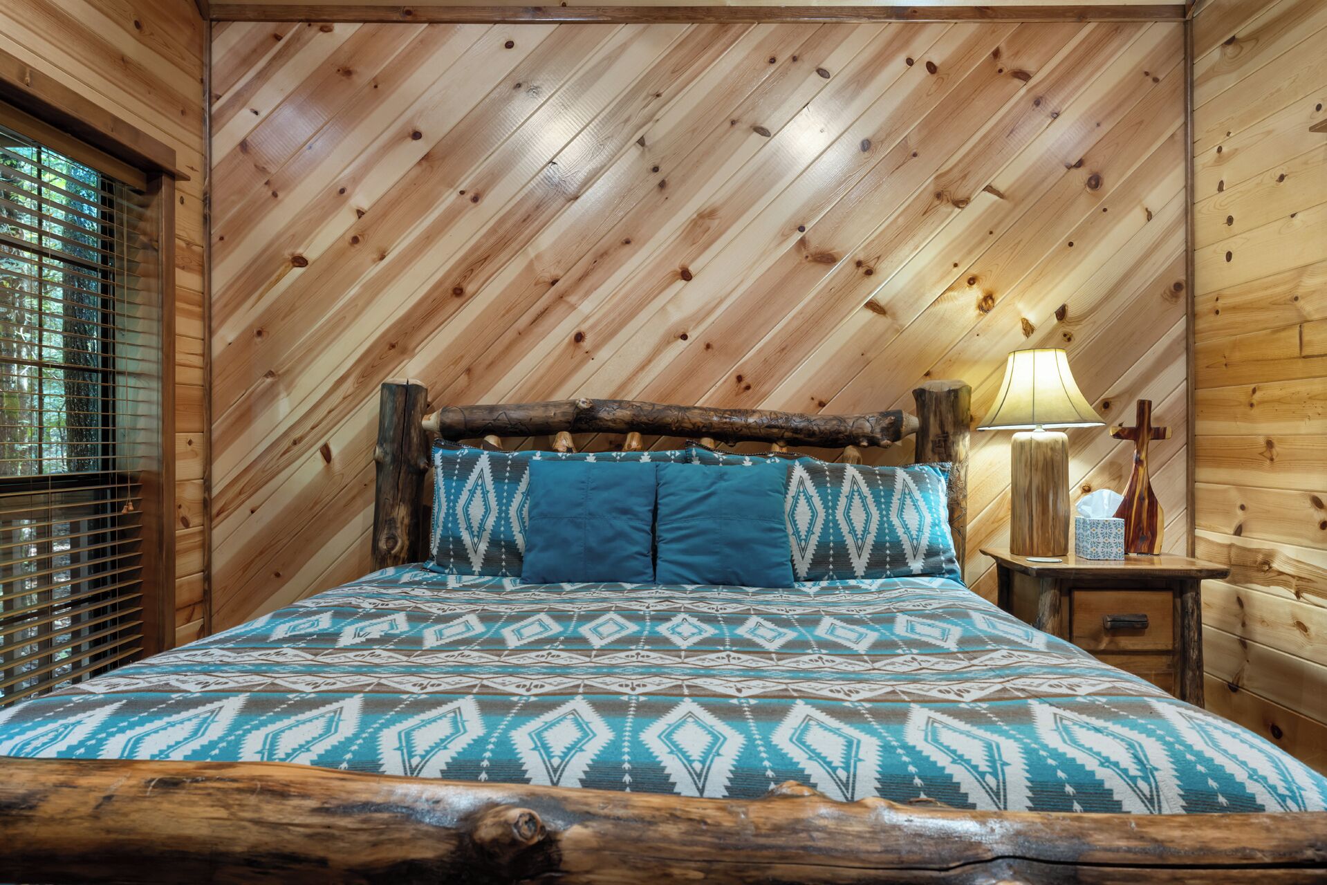 Un dormitorio de cabaña de madera con cama, almohadas, mesita de noche, lámpara y una cruz.