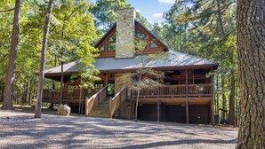 A cabin with a stone chimney, wooden deck, and a gravel driveway surrounded by trees.