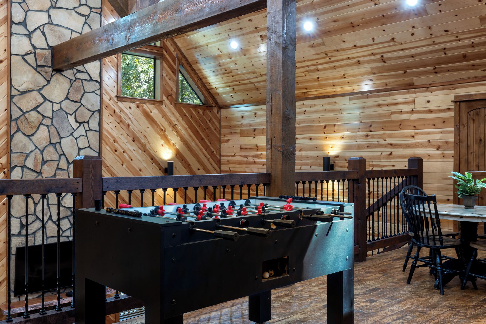 A cozy indoor setting with a foosball table, stone wall, wooden ceiling, and a dining area with chairs and a table.