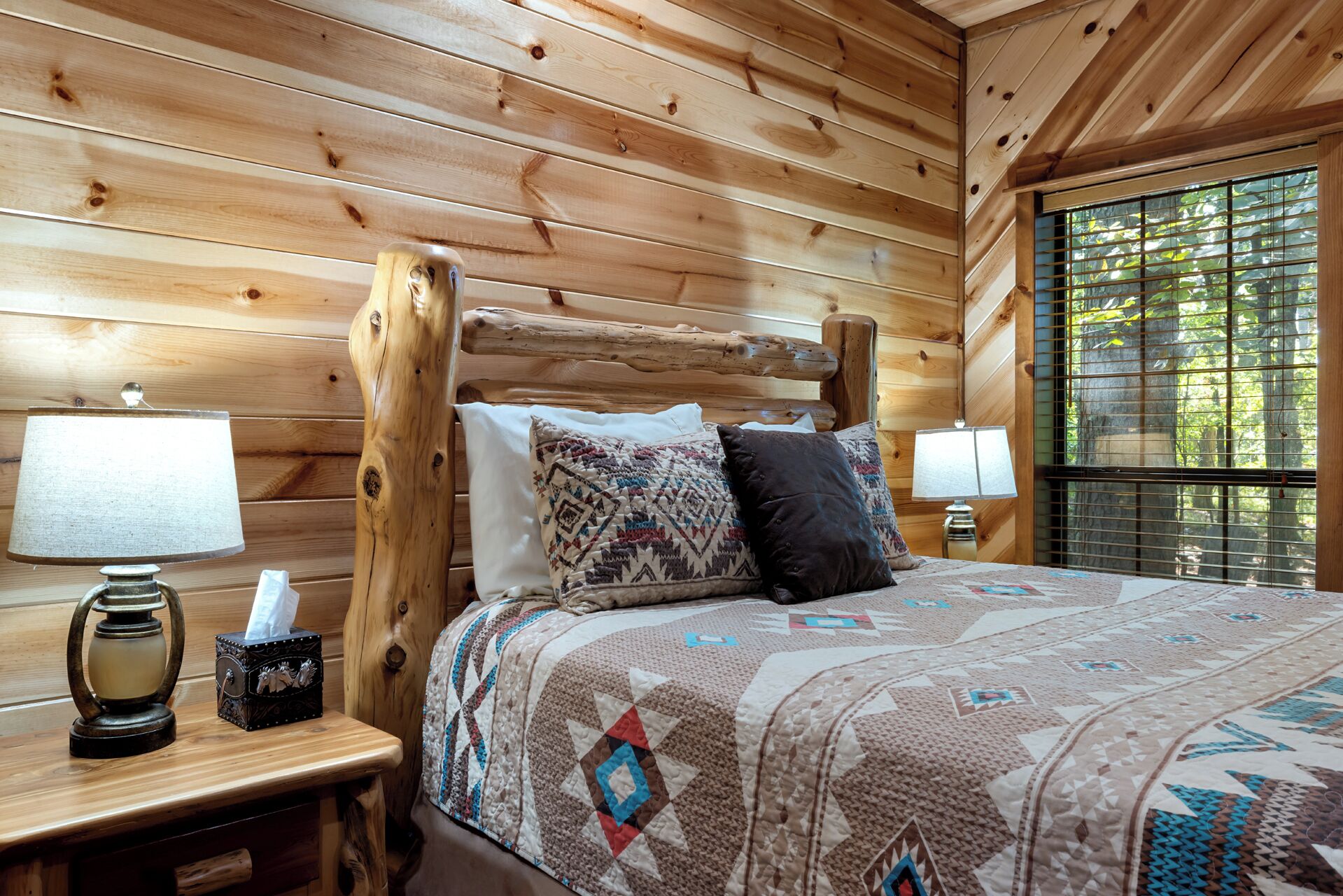 Habitación de cabaña de madera con cama, dos lámparas de noche, mesita de noche con caja de pañuelos y ventana con persianas.