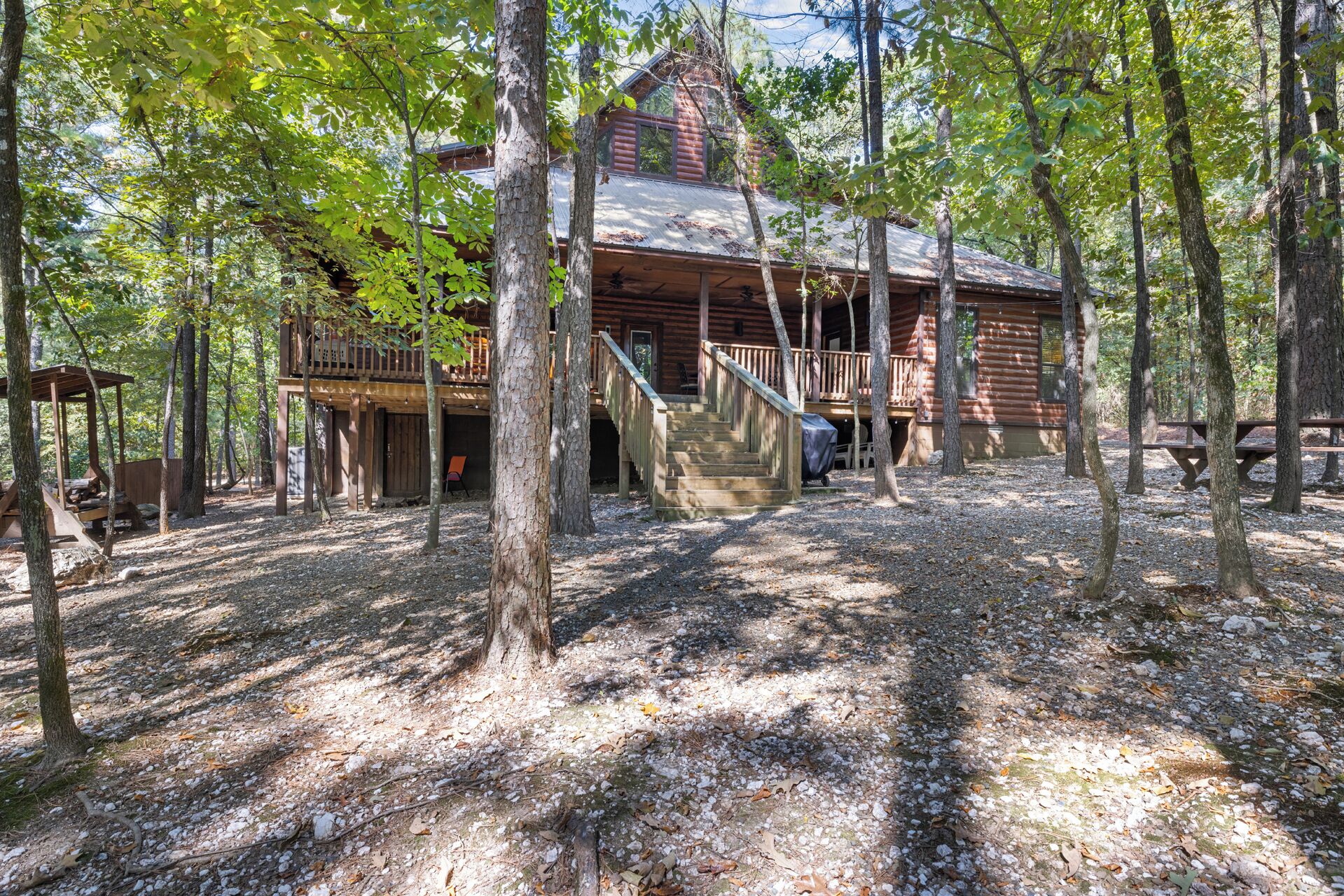 A rustic cabin with a wooden deck, a staircase leading to a porch, and a covered picnic table.