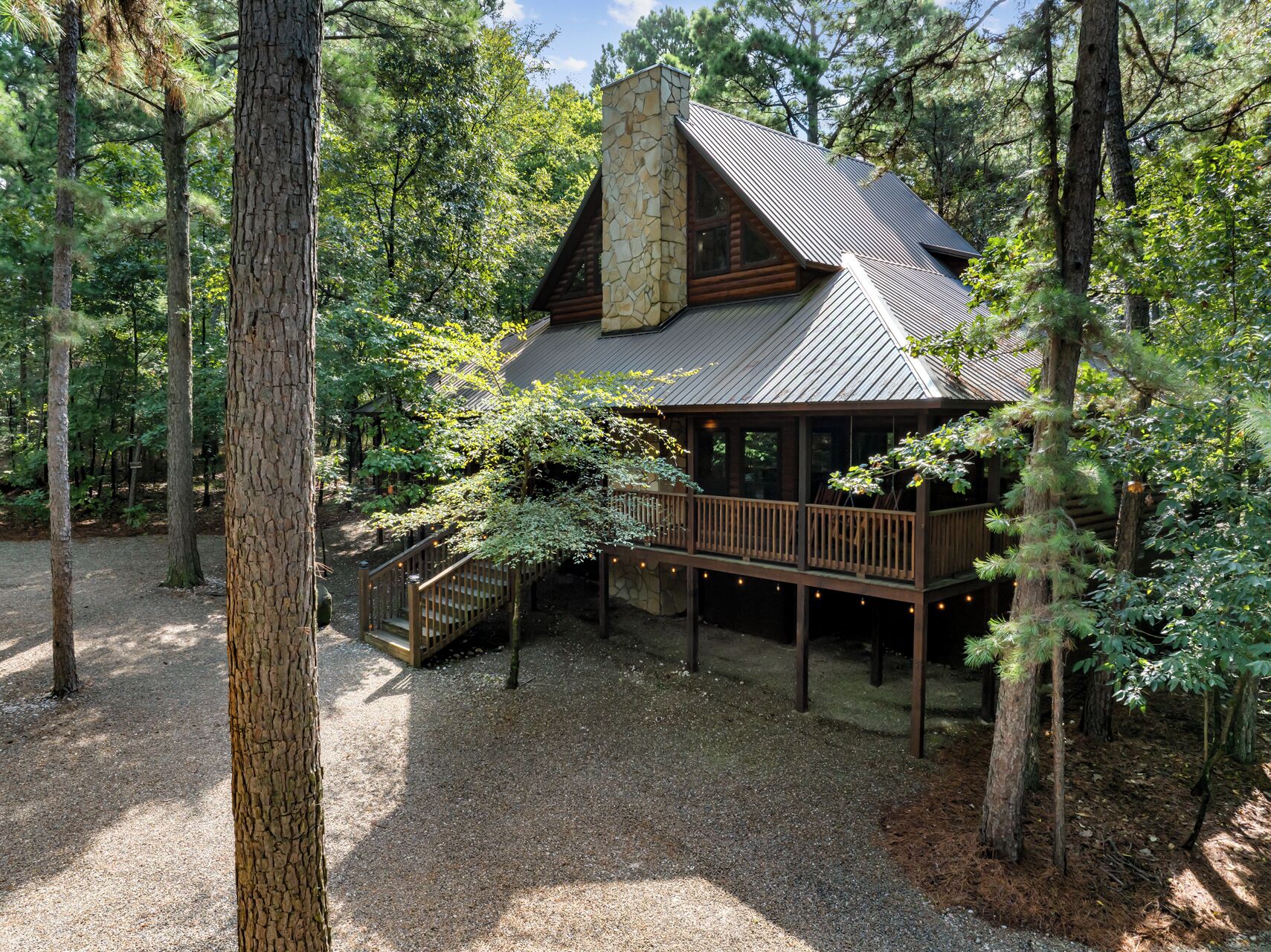 Une cabane avec une cheminée en pierre, une terrasse en bois et entourée d’arbres.