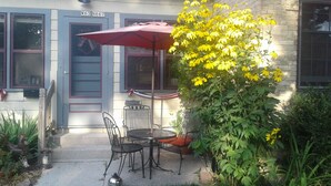 Un espace extérieur aménagé avec des chaises en métal et un parasol rouge, un petit jardin avec des fleurs jaunes et un bâtiment doté d’une porte et de fenêtres bleues.