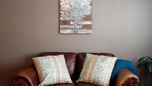 Un canapé en cuir marron agrémenté de coussins à motifs, un tapis blanc et moelleux, et une œuvre d’art représentant un arbre, accrochée au mur.