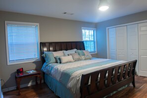 A bedroom with a wooden bed, two windows with blinds, a bedside table with a clock, and a closet with doors.