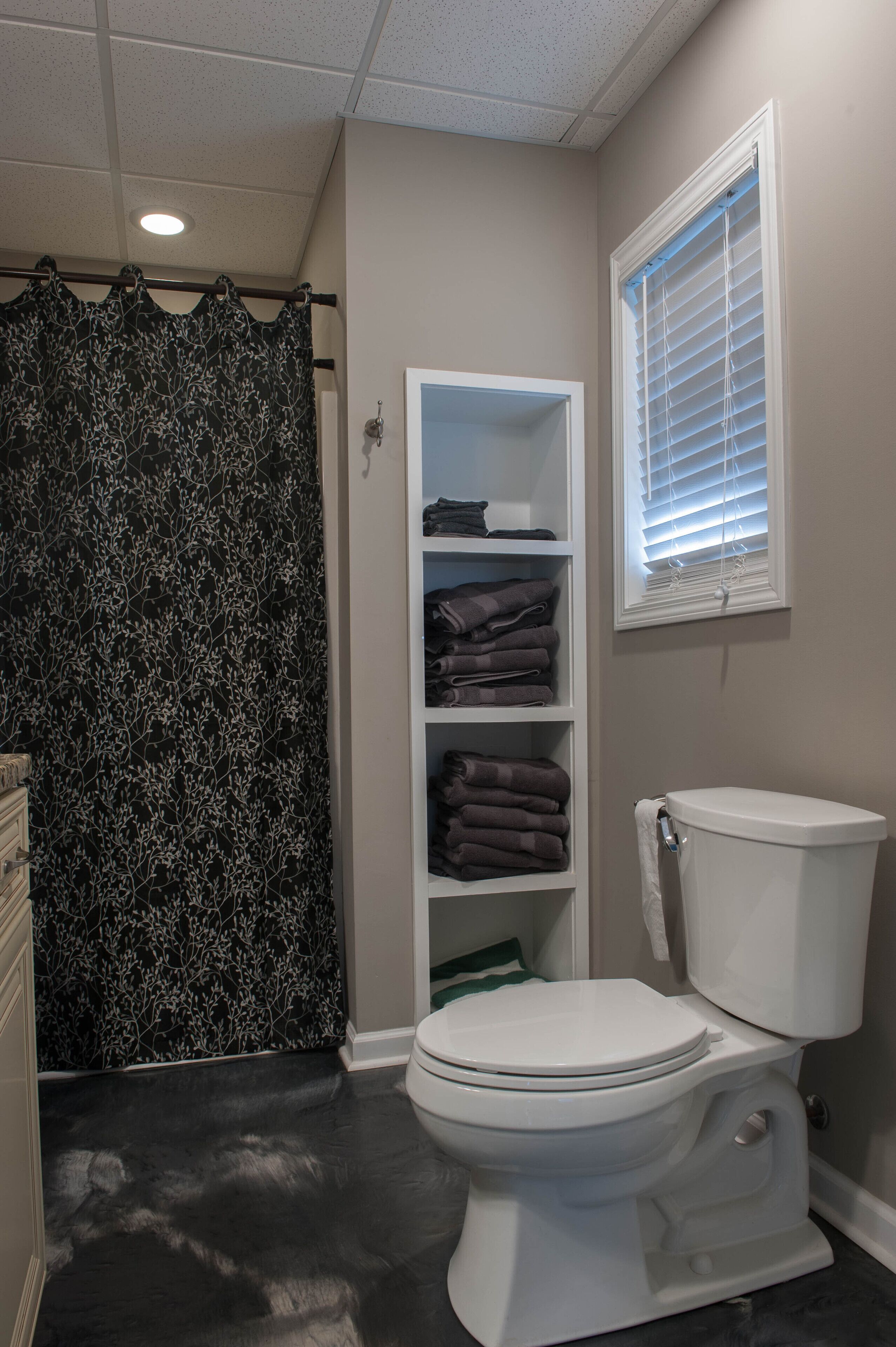 Een badkamer met toilet, een plank met opgevouwen handdoeken, een douchegordijn en een raam met zonwering.