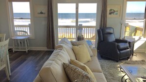 Ett vardagsrum med en beige soffa, en svart läderfåtölj och ett soffbord i glas. Det finns två fönster med utsikt över stranden.