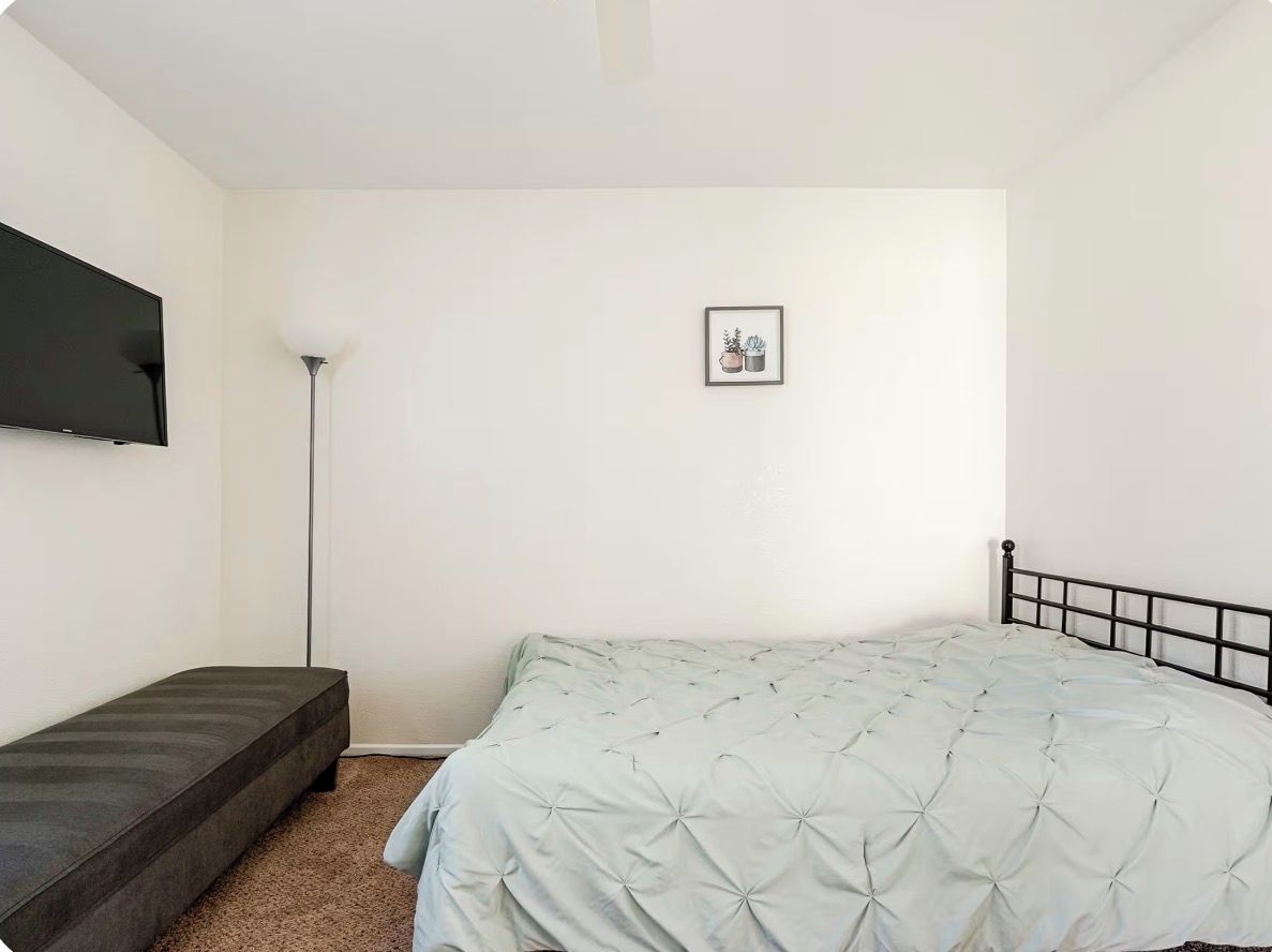 Ein Schlafzimmer mit einem Bett, einer Bank, einer Stehlampe und einem gerahmten Bild an der Wand.