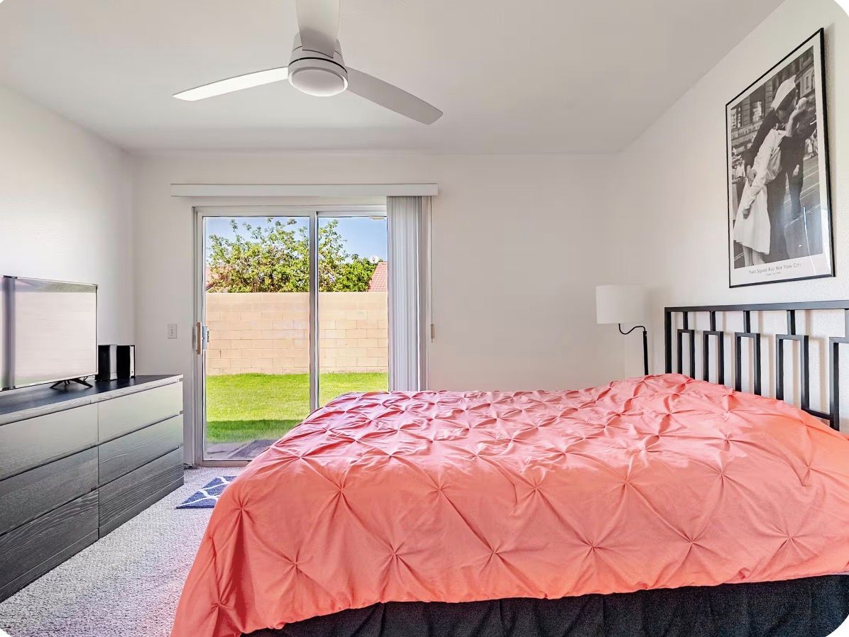 A bedroom with a bed, a ceiling fan, a television, and a sliding glass door leading to a balcony.