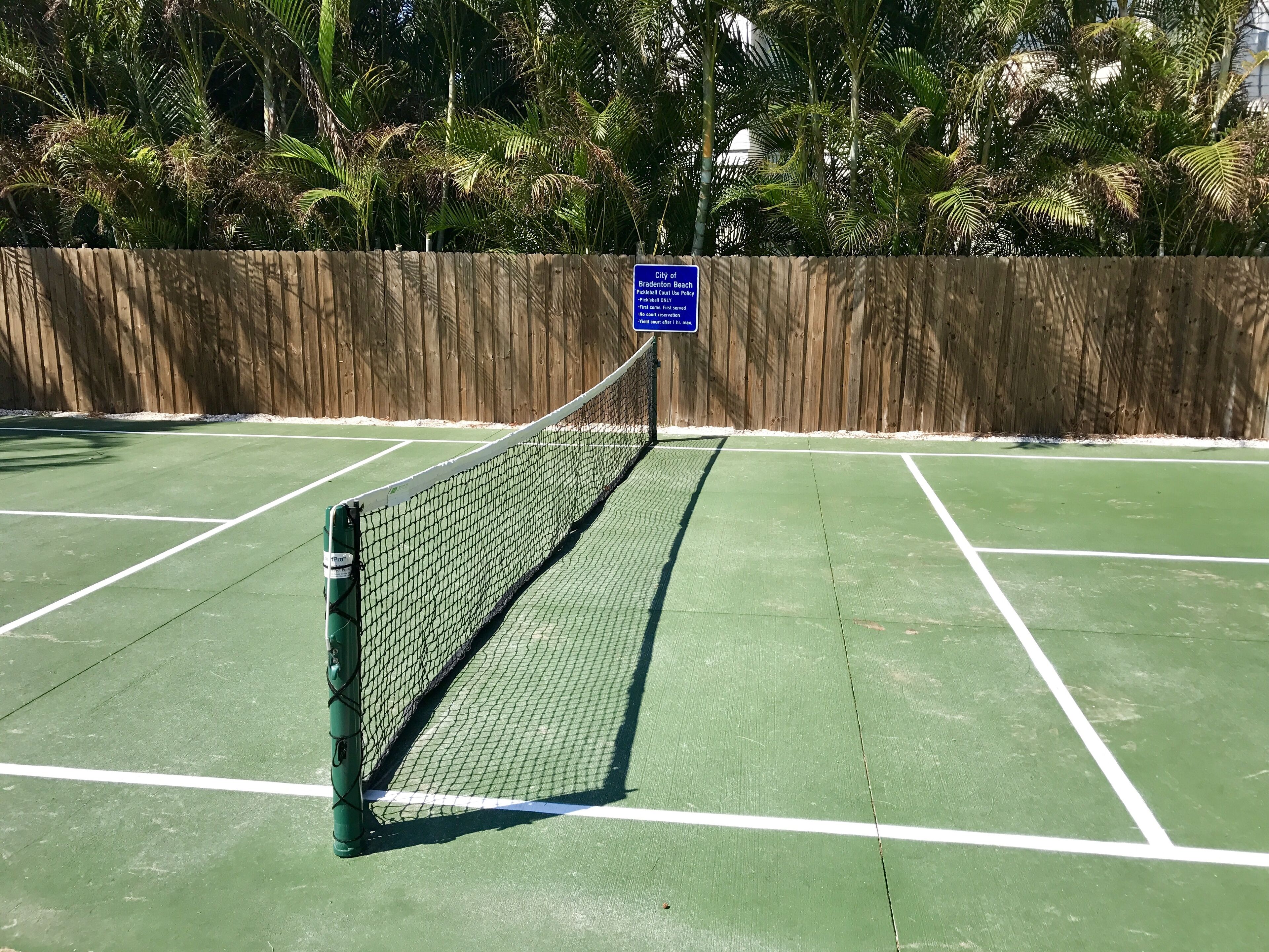 Un court de tennis entouré d’une clôture en bois et muni d’un panneau indiquant qu’il est accessible au public.