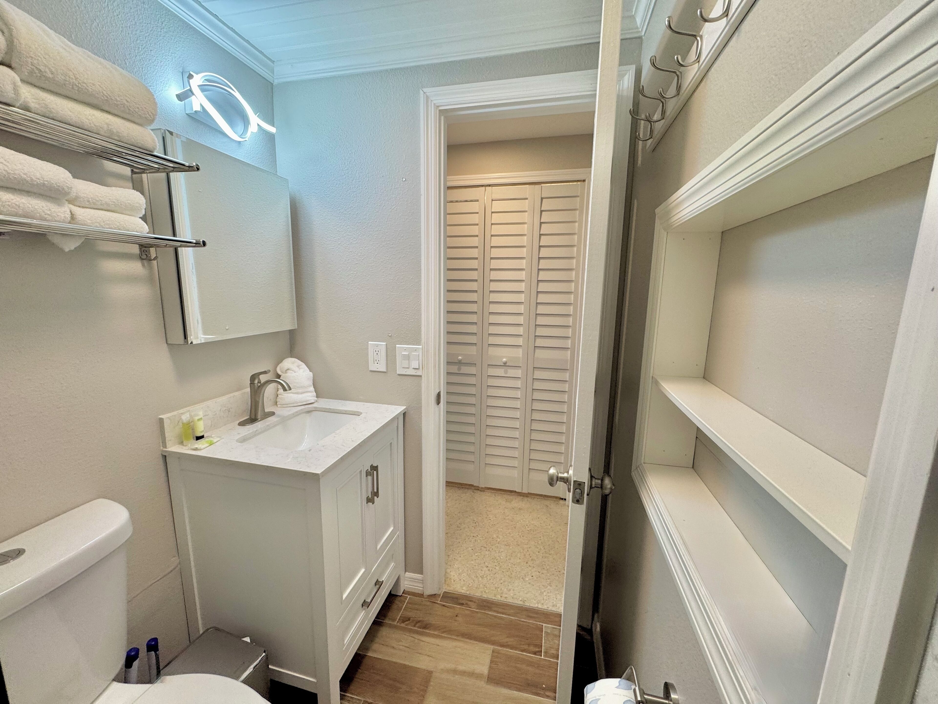 Une salle de bain avec un lavabo blanc, un miroir et un porte-serviettes. On y trouve également des toilettes et une porte qui donne sur une autre pièce.