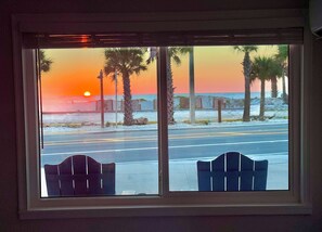 Ein Fenster mit zwei Stühlen, das Blick auf einen Sonnenuntergang über einer Straße mit Palmen und einem Strand bietet.