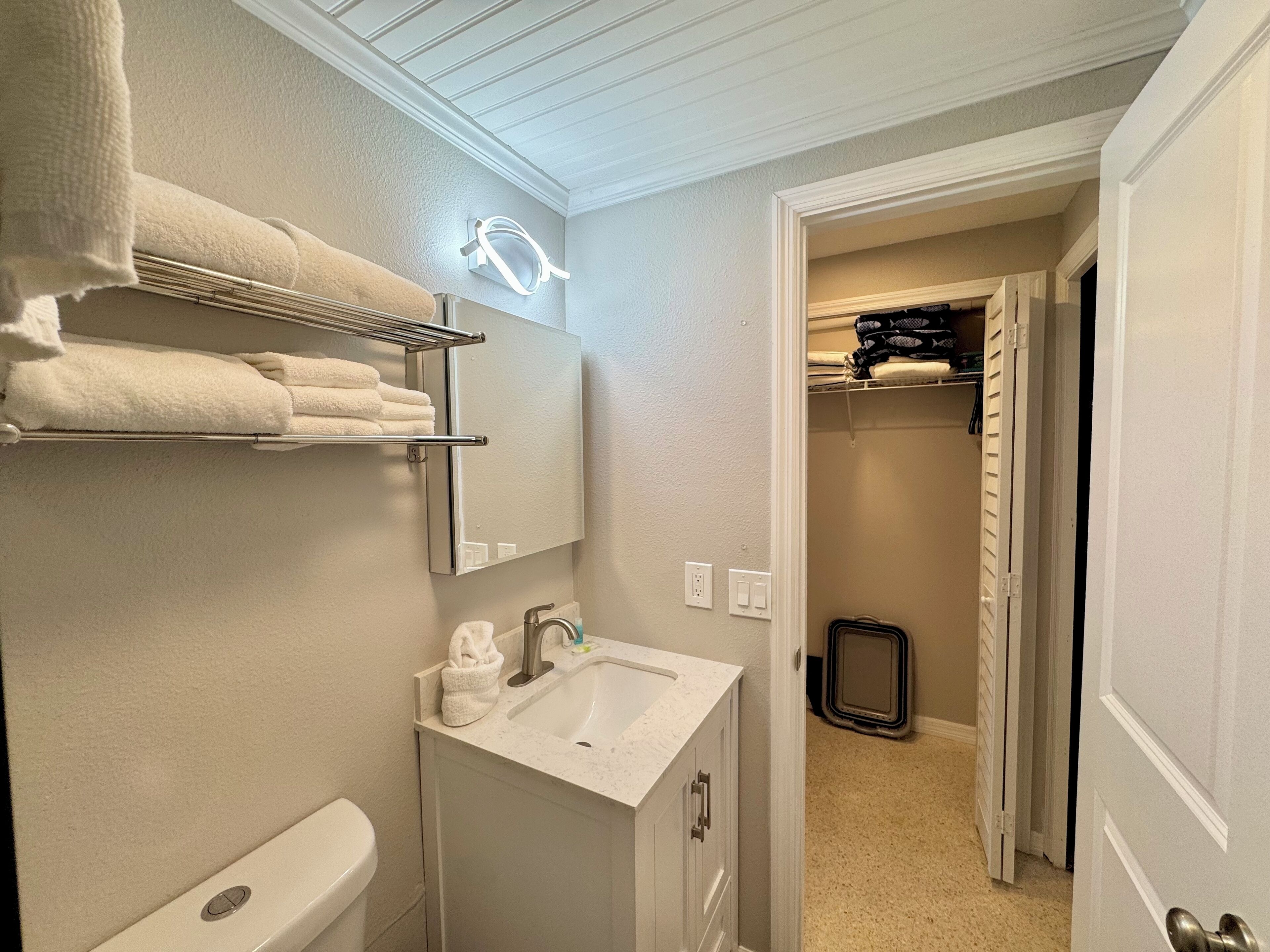A bathroom with a white sink, a towel rack with folded towels, a mirror, and a door leading to a closet.