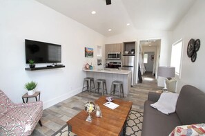 Ein modernes Wohnzimmer mit Flachbildfernseher, Couchtisch und Sofa. Im Hintergrund ist eine Küche mit Bar und Hockern zu sehen.