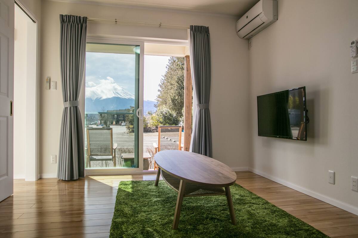 A room with a wooden floor, a green rug, a table, a TV mounted on the wall, and a view of mountains through an open door.